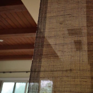 Puede incluir: Una persiana de ventana de bamb&uacute; tejido marr&oacute;n con un patr&oacute;n de rejilla semitransparente. El material tiene una apariencia texturizada natural con bandas verticales m&aacute;s oscuras en los bordes, permitiendo que la luz filtre a trav&eacute;s.