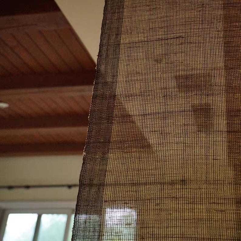 Puede incluir: Una persiana de ventana de bamb&uacute; tejido marr&oacute;n con un patr&oacute;n de rejilla semitransparente. El material tiene una apariencia texturizada natural con bandas verticales m&aacute;s oscuras en los bordes, permitiendo que la luz filtre a trav&eacute;s.