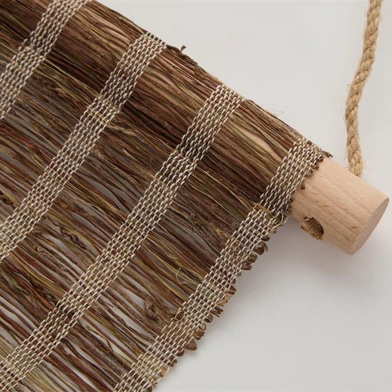 Puede incluir: Un tapiz de pared hecho de fibras naturales marrones tejidas con hilo beige claro, sujeto a una varilla cil&iacute;ndrica de madera clara. Una cuerda gruesa y trenzada de color beige pasa por un orificio en el extremo de la varilla de madera.