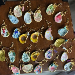 May include: A selection of decorative keyrings with gold-coloured hardware. The keyrings feature painted designs, including floral patterns, sunflowers, and abstract art, with colourful tassels. The keyrings are arranged on a wooden surface.
