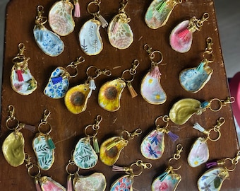 May include: A collection of decorative keychains with gold-colored hardware. The keychains feature painted designs, including floral patterns, sunflowers, and abstract art, with colorful tassels. The keychains are arranged on a wooden surface.