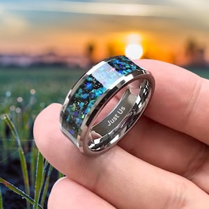 Puede incluir: Un anillo plateado con una banda de material azul y verde iridiscente. El anillo tiene el texto grabado "Just Us" en el interior. El anillo se muestra sobre un fondo borroso de campo y puesta de sol.