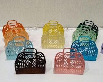 Può includere: Una collezione di borse tote colorate, in plastica, a trama aperta, in varie tonalità tra cui viola, arancione, giallo, verde, blu, nero e rosa. Ogni borsa ha un manico e un design a forma di cestino, adatta per trasportare oggetti.