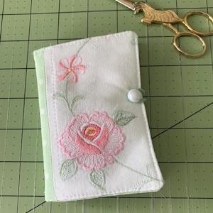 May include: A white fabric journal cover with a pink rose and flower embroidery design. The cover has a light green border with white polka dots and a button closure. Gold scissors are in the background.