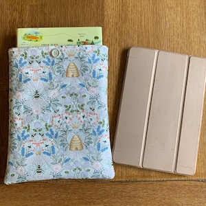 May include: A white fabric book sleeve with a bee and honeycomb pattern. The sleeve is filled with a book with a map on the cover. A beige tablet case is next to the sleeve.