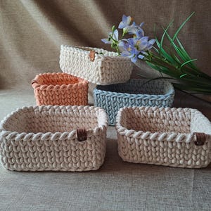 Könnte beinhalten: Fünf rechteckige Häkelkörbe in Creme, Pfirsich und Blau. Jeder Korb hat einen geflochtenen Rand und ein kleines Lederetikett. Ein Strauß blauer und grüner Blumen liegt daneben. Handgefertigt.
