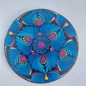 May include: A circular disc featuring a hand-painted mandala design on a black background. The artwork consists of symmetrical patterns in shades of blue, purple, and gold, utilizing dot art techniques to create intricate floral-like shapes.