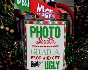 May include: A clear glass jar with a green, red, and white plaid pattern label that says "Photo Booth Grab a Prop and Get Ugly". The jar is filled with festive props for a photo booth.