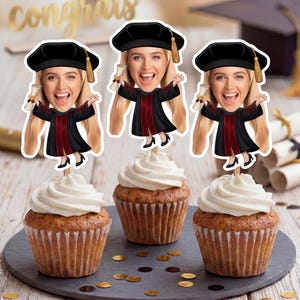 May include: Three cupcakes topped with white frosting, each adorned with a graduation-themed topper. The toppers feature a smiling person in a graduation cap and gown, holding a diploma. The cupcakes sit on a dark gray plate with gold confetti.