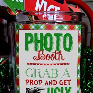 May include: A clear glass jar with a red and green plaid label that says "Photo Booth Grab a Prop and Get Ugly". The jar is filled with various props for a photo booth, including a green and white sign that says "NAUGHTY" and a red sign that says "NICE".