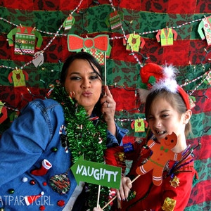 May include: Two people wearing festive Christmas jumpers pose for a photo in front of a red and green patterned backdrop. The woman on the left is holding a prop with a red bow and the text "NAUGHTY". The girl on the right is holding a gingerbread man prop.
