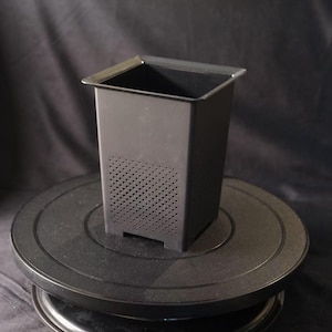 Peut inclure: Un récipient rectangulaire noir avec un côté perforé. Le récipient a un dessus carré et repose sur une base circulaire noire. Le fond est un tissu sombre et texturé.