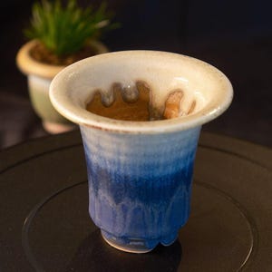 May include: A ceramic cup with a flared rim and a blue and white glaze. The cup has a unique shape and a textured surface. A small potted plant is in the background.
