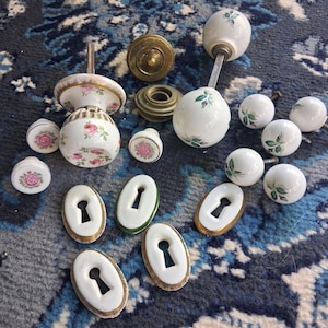 May include: A collection of vintage door hardware, including white porcelain doorknobs with pink rose and green leaf designs, brass fixtures, and keyhole covers with gold trim. The hardware is arranged on a blue and white patterned surface.