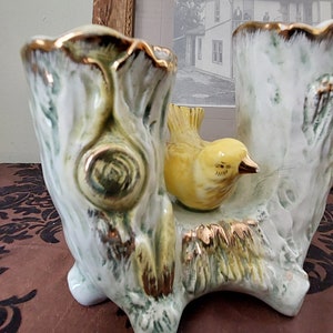May include: A ceramic planter shaped like a tree trunk with two openings for plants. A small yellow bird figurine is perched on the trunk.