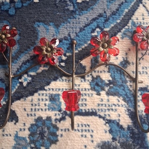 May include: A decorative wall sconce with a floral design. The sconce features four red flower-shaped accents with bronze-colored centers. The sconce has a dark metal frame and red, faceted candle holders.