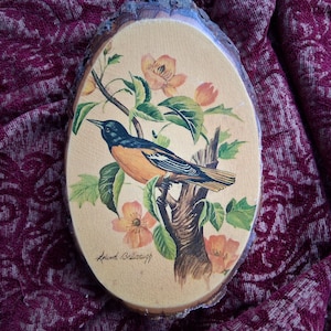 May include: Oval wooden plaque featuring a hand-painted bird perched on a tree branch with orange flowers and green leaves. The bird has black, orange, and blue plumage. The artwork is signed in the lower left corner.