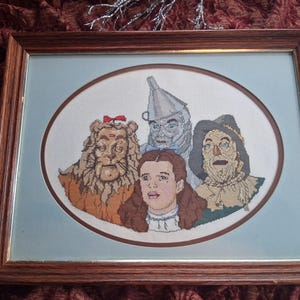 May include: Framed cross-stitch artwork featuring characters from The Wizard of Oz. The artwork depicts the Lion, Tin Man, Dorothy, and Scarecrow in an oval frame. The frame is made of brown wood.