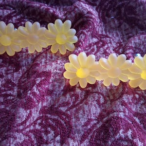 May include: Two pairs of daisy-shaped earrings. Each earring features three yellow daisy flowers with a frosted appearance. The earrings are set against a textured, patterned, purple background.