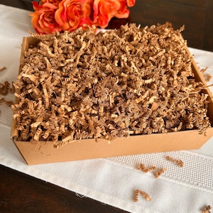 May include: A rectangular brown cardboard box filled with shredded brown paper. The box is sitting on a white surface. Orange roses are in the background.