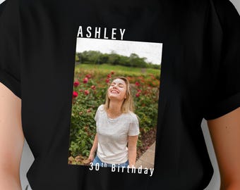 May include: Black t-shirt with white text that says "Ashley 30th Birthday". The shirt features a photo of a woman smiling in a field of flowers.