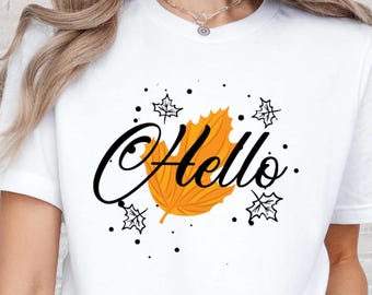 May include: White t-shirt featuring the word "Hello" in black script, with an orange leaf and decorative black accents. The shirt is a short-sleeved crew neck.