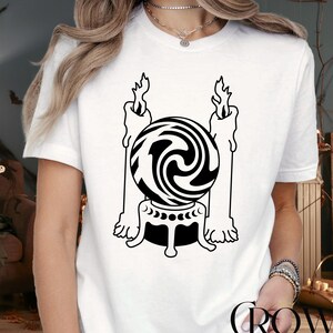 May include: White t-shirt featuring a black and white graphic of a crystal ball between two candles. The crystal ball is on a stand with a crescent moon design. The shirt has the text "CROWN CUSTOM SHIRT" in a circular design.