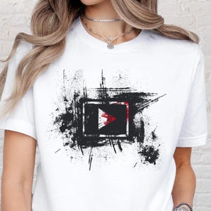 May include: White t-shirt with a black and red graphic. The design features a black square containing a red and white arrow shape, set against a background of black paint strokes.