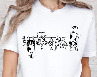 May include: White t-shirt featuring a black and white illustration of several cartoon cats hanging from a clothesline. The cats have various patterns and are accompanied by small fish and socks. The shirt is a casual, everyday wear item.