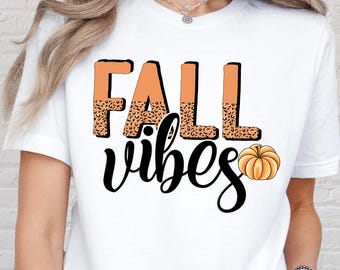 May include: White t-shirt with the words "FALL vibes" in a fall-themed design. The word "FALL" is in orange with a leopard print pattern, and "vibes" is in a black script font. A small orange pumpkin graphic is next to the word "vibes".