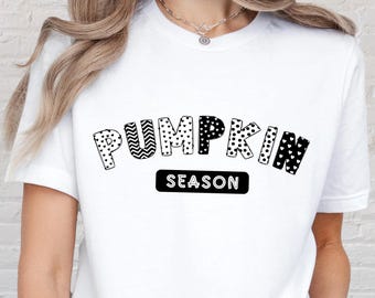 May include: White t-shirt with the text "PUMPKIN SEASON" in a black, patterned font. The word "PUMPKIN" is arched, and "SEASON" is in a black rectangle. The shirt has short sleeves.