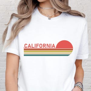 May include: White t-shirt with the word "CALIFORNIA" in red, above a retro sunset design with horizontal stripes in yellow, orange, and green. The shirt is a classic crew neck style.