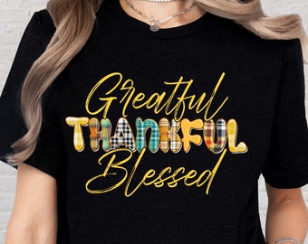 May include: Black t-shirt with the words "Grateful, Thankful, Blessed" in a mix of yellow script and plaid block letters. The shirt is a crew neck and has short sleeves.