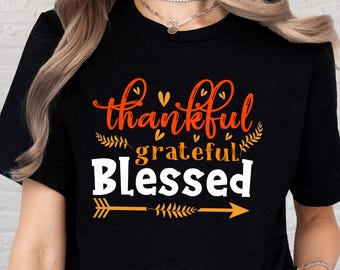 May include: Black t-shirt with the words "thankful, grateful, blessed" in orange and white script. The design includes decorative arrows and leaf accents, perfect for Thanksgiving or autumn.