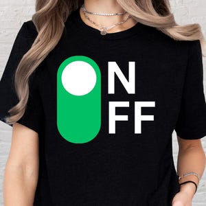 May include: Black t-shirt with a green and white graphic. The graphic features a green rectangle with a white circle and the letters "ON" and "FF" in white. The shirt is a crew neck.