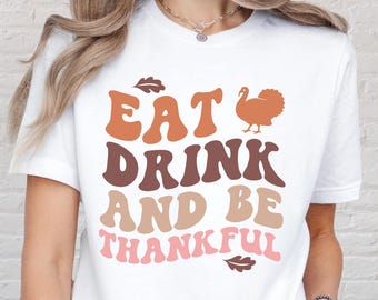 May include: White t-shirt with the text "EAT DRINK AND BE THANKFUL" in retro-style lettering. The design includes a turkey illustration and leaf accents in shades of brown and pink. A casual, festive top.