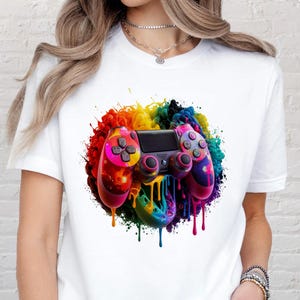 May include: White t-shirt featuring a colorful, artistic rendering of a video game controller. The controller is depicted with vibrant rainbow hues and paint drip effects, set against a white background. The design is centered on the shirt.