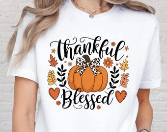 May include: White t-shirt featuring a fall-themed graphic. The design includes an orange pumpkin with a leopard print bow, surrounded by leaves, flowers, and the words "thankful" and "blessed" in black script.