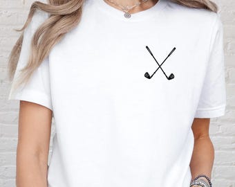 May include: White t-shirt featuring a small, black graphic of two crossed golf clubs. The design is centered on the chest. The shirt is made of a soft, likely cotton material. The overall style is casual and sporty.