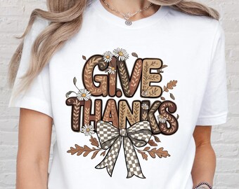 May include: White t-shirt featuring the words "GIVE THANKS" in a decorative, fall-themed design. The design includes a checkered bow, daisies, and autumn leaves in shades of brown and beige.