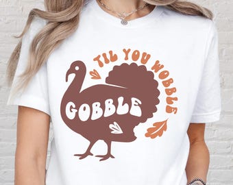 May include: White t-shirt with a brown turkey graphic and the text "TIL YOU WOBBLE GOBBLE". The turkey design includes a heart and two orange leaves. A casual, festive top for Thanksgiving.