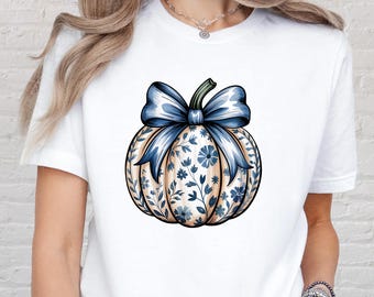 May include: White t-shirt featuring a detailed illustration of a pumpkin with a blue bow and floral pattern. The pumpkin design incorporates blue and cream colors, creating a seasonal and decorative aesthetic.