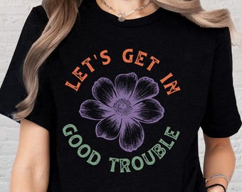 May include: Black t-shirt with the text "LET'S GET IN GOOD TROUBLE" in orange and green. A purple flower graphic is centered within the text. The shirt is a crew neck style.
