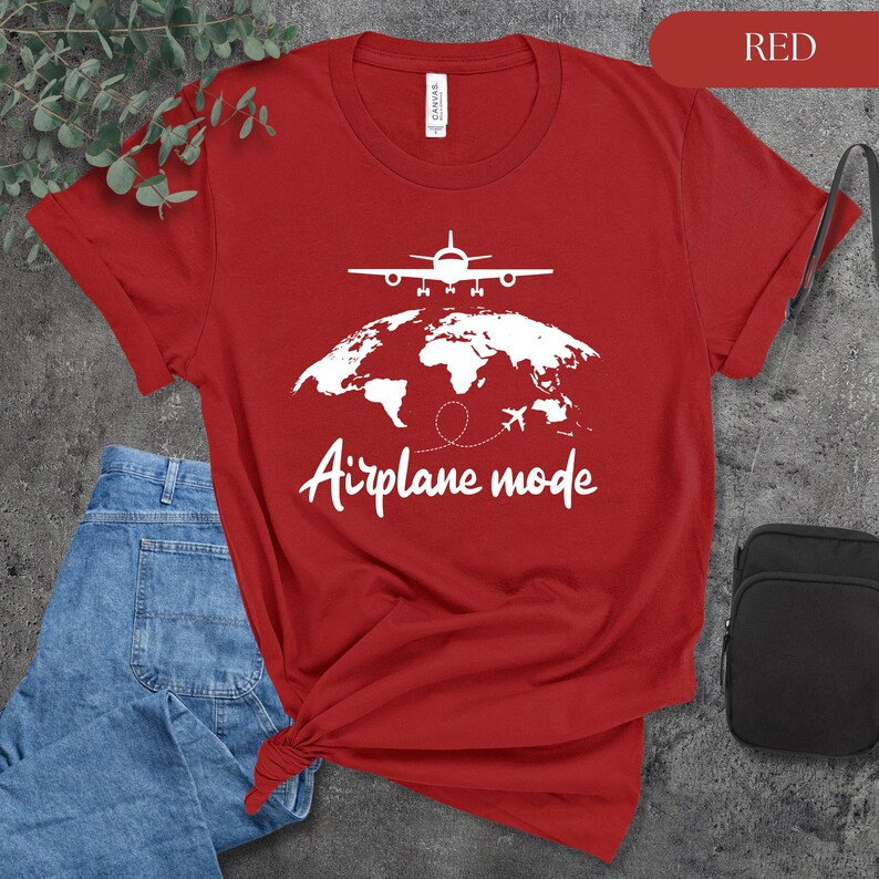 May include: A red t-shirt with a white graphic of an airplane above a world map and the words "Airplane mode". The shirt is paired with blue jeans and a black bag.