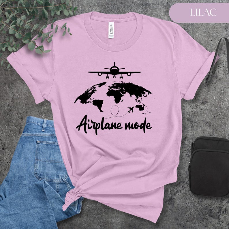 May include: Lilac-colored t-shirt with a black graphic of an airplane, a world map, and the words "Airplane mode." The shirt is made of soft material and is suitable for casual wear. The shirt is paired with blue jeans.