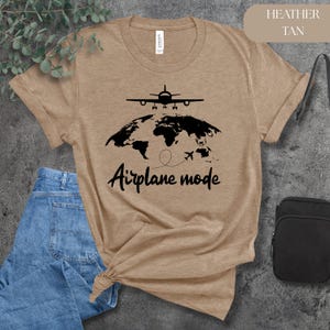 May include: Heather tan t-shirt with a black graphic of an airplane above a world map and the words "Airplane mode". The shirt is paired with blue jeans and a black crossbody bag.