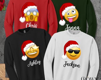 May include: Four crewneck sweatshirts in red, green, black, and white. Each features a different emoji wearing a Santa hat, with names Chris, Jenna, Ashley, and Jackson below. The sweatshirts are a festive holiday theme.