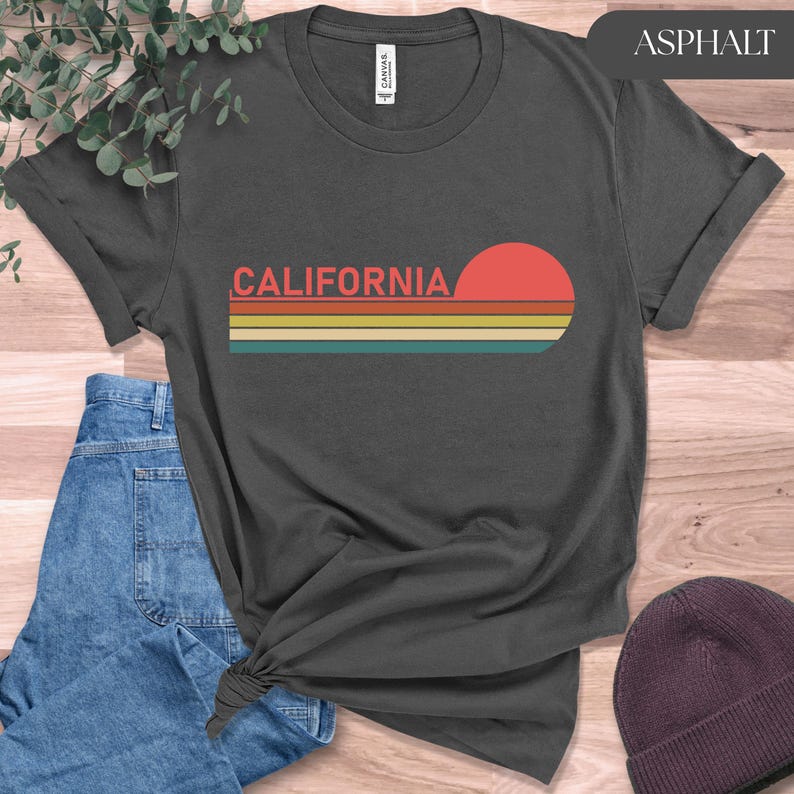May include: Asphalt-colored t-shirt featuring the word "CALIFORNIA" in coral lettering, with a retro sunset design in red, yellow, and teal stripes. The shirt is knotted at the waist and paired with blue jeans and a maroon beanie.
