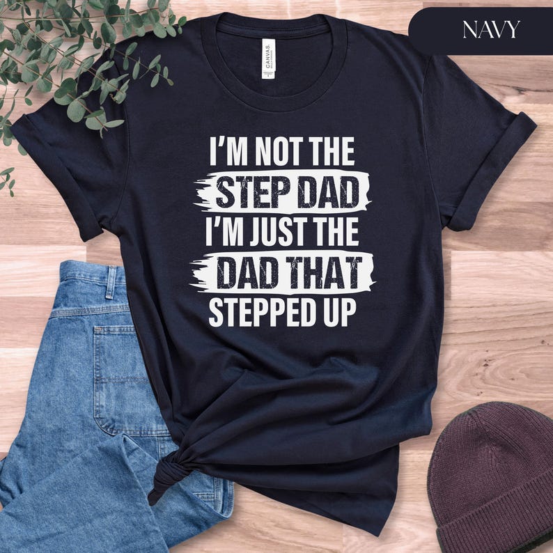 Puede incluir: Camiseta azul marino con el texto blanco "I'M NOT THE STEP DAD I'M JUST THE DAD THAT STEPPED UP". La camiseta est&aacute; doblada, con unos vaqueros azules y un gorro marr&oacute;n en el fondo. La palabra "NAVY" est&aacute; en la esquina superior derecha.