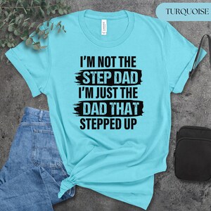 Puede incluir: Camiseta turquesa con el texto negro "I'M NOT THE STEP DAD I'M JUST THE DAD THAT STEPPED UP". La camiseta es de color azul claro y est&aacute; doblada en la parte inferior. Se ven unos vaqueros azules a la izquierda.